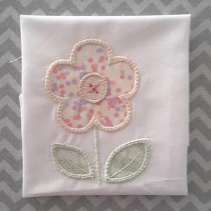May include: A white fabric with a pink and white embroidered flower with a green stem and leaves.