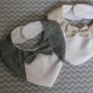 May include: Two white bibs with a white fleece lining and a bow tie design. One bib has a green and white polka dot bow tie and the other has a beige and white polka dot bow tie.