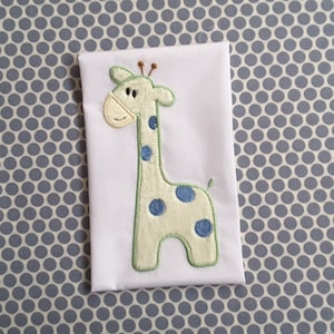 May include: White fabric with an embroidered giraffe design. The giraffe is light green with blue spots and a green outline. The background fabric is white, and the giraffe has brown antennae and a tan face. The background is a blue and white polka dot pattern.