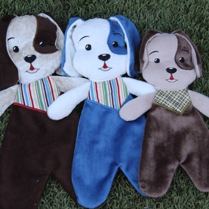 Op de afbeelding: Drie pluche hondenspeelgoed met verschillende gekleurde outfits. De eerste hond is bruin en tan met een gestreepte outfit. De tweede hond is wit met een blauw-wit gestreepte outfit en een blauwe overall. De derde hond is bruin met een geruite outfit.