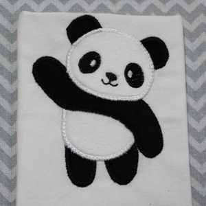 Può includere: Un tessuto bianco con un orso panda ricamato in bianco e nero. Il panda è in piedi sulle zampe posteriori con il braccio sinistro alzato in un gesto di saluto. Il panda ha un naso nero e occhi neri.