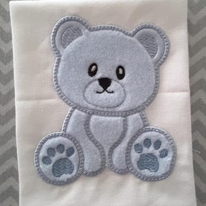 Puede incluir: Una aplicación de osito de peluche bordada en azul claro sobre un fondo de tela blanca.