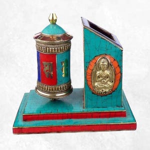 May include: A decorative turquoise and red desk organizer with a prayer wheel and a Buddha statue. The prayer wheel is cylindrical with gold accents and colorful panels. The Buddha is in a gold-colored medallion.