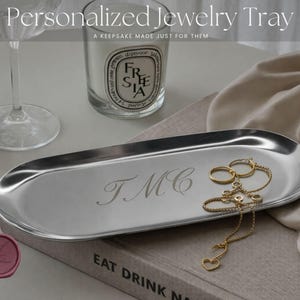 Puede incluir: Una bandeja de joyería ovalada plateada con iniciales grabadas, que contiene anillos y un collar de oro. La bandeja está sobre un libro con el texto "EAT DRINK NA". La imagen también incluye una vela y una copa de vino. El texto de la imagen dice "Personalized Jewelry Tray" y "A keepsake made just for them."