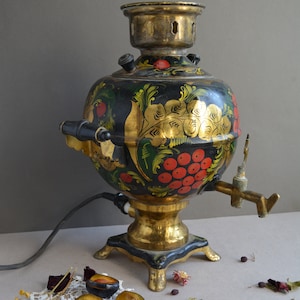 May include: A traditional Russian samovar with intricate floral and berry designs painted in gold, red, and green on a black background. The samovar has a brass spout and a brass base with three legs.
