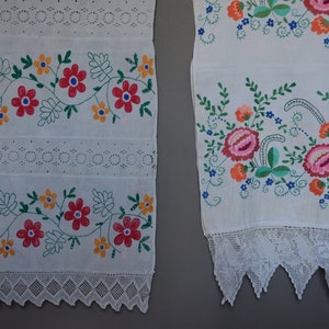 May include: Two white embroidered fabric panels with floral designs. The panels have a lace trim at the bottom. The floral designs are in shades of red, pink, orange, yellow, green, and blue.