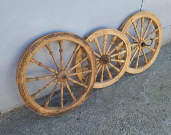 May include: Three antique wooden wagon wheels leaning against a light blue wall. The wheels are made of light brown wood, with spokes radiating from a central hub. The wheel on the left shows signs of age and wear, with darker areas.