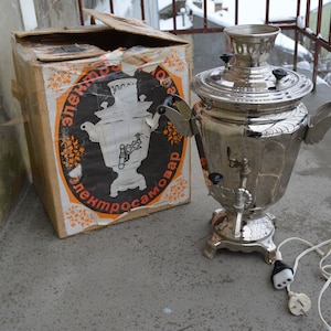 May include: A silver electric samovar with a black and orange box. The box has the text "Электросамовар" printed on it.