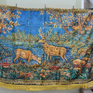 May include: A blue tapestry with a forest scene featuring deer, trees, and a lake. The tapestry has a gold fringe and a black border with a floral pattern. The deer are brown and the trees have gold leaves.