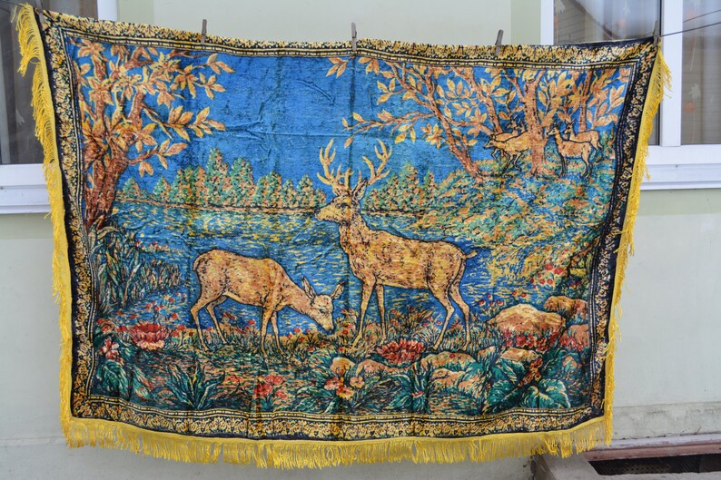 May include: A blue tapestry with a forest scene featuring deer, trees, and a lake. The tapestry has a gold fringe and a black border with a floral pattern. The deer are brown and the trees have gold leaves.