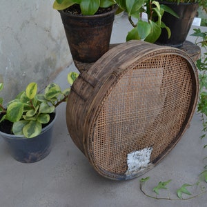 Puede incluir: Un tamiz redondo de madera tejido con un acabado marrón natural. El tamiz está sentado en una mesa con una planta en maceta en el fondo.