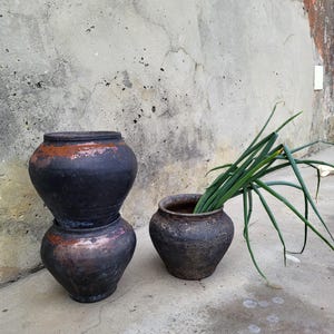 May include: Three dark, rustic ceramic pots with a weathered appearance. Two are stacked, and the third holds green onions. The pots have a dark, matte finish with reddish-brown accents. The scene is set against a textured, grey concrete wall.