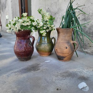Puede incluir: Tres jarras de cerámica rústicas de varios tamaños y colores. La jarra de la izquierda es de color burdeos oscuro, la del centro es verde y la de la derecha es marrón. Cada jarra tiene un asa y está llena de vegetación y flores blancas.