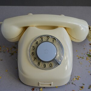 Puede incluir: Un tel&eacute;fono de disco rotatorio vintage con un cuerpo de color crema y un dial negro. El tel&eacute;fono tiene un cable conectado al lateral.