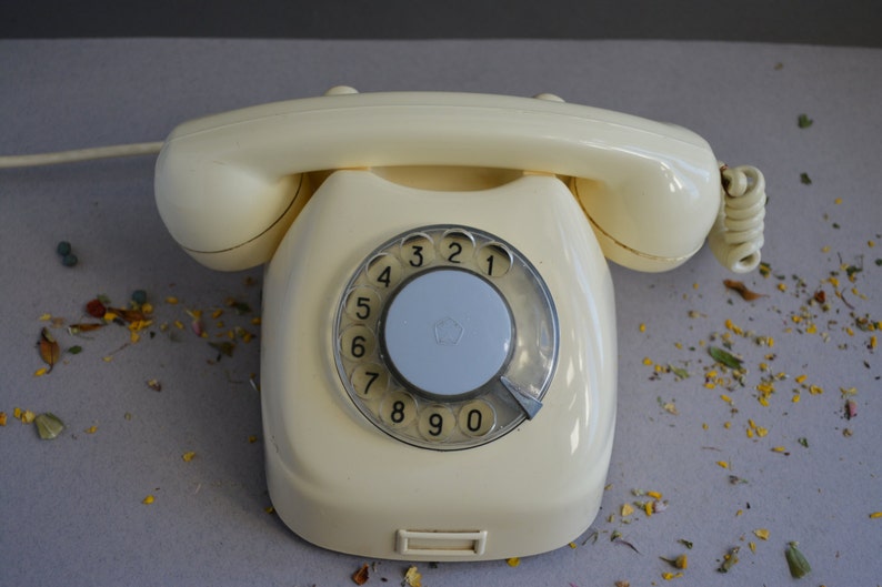 Puede incluir: Un tel&eacute;fono de disco rotatorio vintage con un cuerpo de color crema y un dial negro. El tel&eacute;fono tiene un cable conectado al lateral.