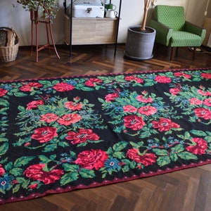 May include: A black and red floral patterned rug with a fringed edge. The rug has a traditional design with pink and green flowers on a black background.