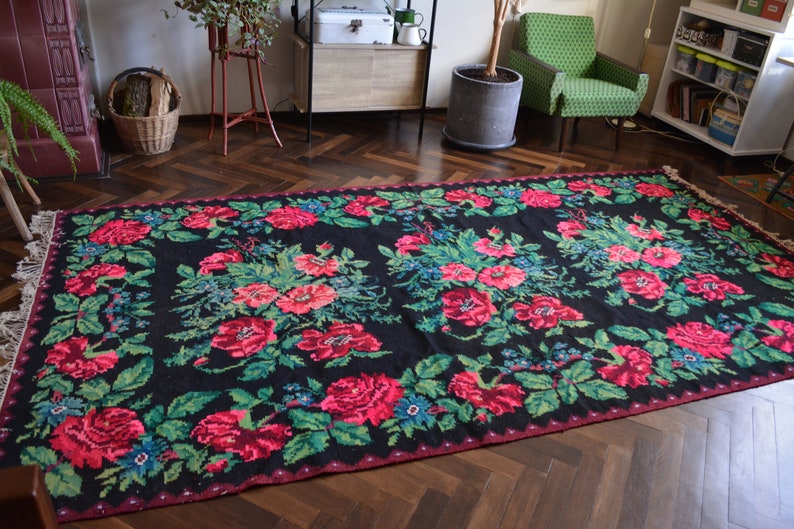 May include: A black and red floral patterned rug with a fringed edge. The rug has a traditional design with pink and green flowers on a black background.