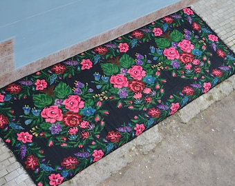 May include: Black rectangular rug with a floral design. The rug has a border of pink, red, and blue flowers with green leaves. It is placed on a concrete surface.
