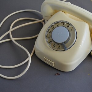 Puede incluir: Un tel&eacute;fono de disco vintage con cuerpo de color crema y disco gris. El tel&eacute;fono tiene un cable en espiral y un cable separado con un enchufe.