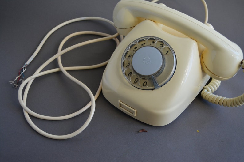 Puede incluir: Un tel&eacute;fono de disco vintage con cuerpo de color crema y disco gris. El tel&eacute;fono tiene un cable en espiral y un cable separado con un enchufe.