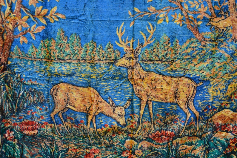 May include: A tapestry depicting two deer in a forest scene. The deer are golden brown with antlers, set against a blue and green backdrop of trees, water, and foliage. The artwork has a vintage, textured appearance.