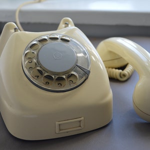 Puede incluir: Un tel&eacute;fono de disco vintage con un cuerpo de color crema y un disco rotatorio negro. El tel&eacute;fono tiene un auricular separado con un cuerpo de color crema.