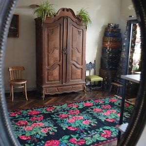 May include: A vintage wooden armoire with two doors and a drawer, standing in a room with a patterned rug and a stack of suitcases. The armoire has ornate carvings and a dark wood finish.