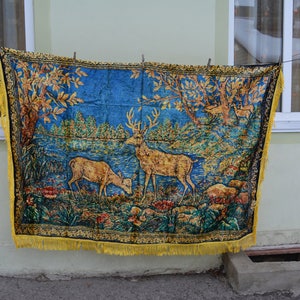 May include: A tapestry featuring a forest scene with two deer, trees, and flowers, in shades of blue, gold, and green. The tapestry has a yellow fringe along the bottom edge and is hanging on a clothesline.