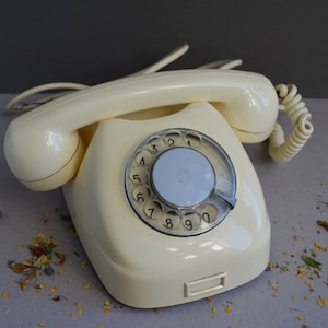 Puede incluir: Un tel&eacute;fono de disco vintage de color crema con un dial y n&uacute;meros negros. El tel&eacute;fono tiene un cable en espiral y un auricular.