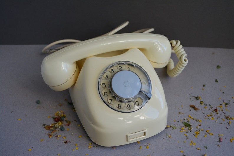Puede incluir: Un tel&eacute;fono de disco vintage de color crema con un dial y n&uacute;meros negros. El tel&eacute;fono tiene un cable en espiral y un auricular.