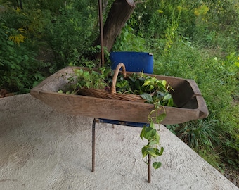 May include: A rustic, wooden trough filled with greenery and a woven basket. The trough is supported by a metal frame. The scene is set outdoors with lush foliage in the background.