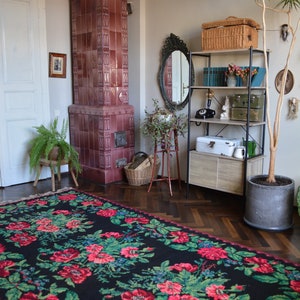 May include: A black and red floral rug with green leaves, a tiled wood-burning stove, a wooden shelf with a wicker basket, a mirror, and a potted plant in a room with hardwood floors.