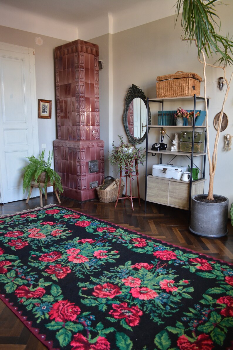 May include: A black and red floral rug with green leaves, a tiled wood-burning stove, a wooden shelf with a wicker basket, a mirror, and a potted plant in a room with hardwood floors.