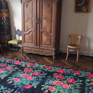 May include: A black and red floral patterned rug with green leaves. The rug is in front of a wooden armoire with two doors and a wooden chair.