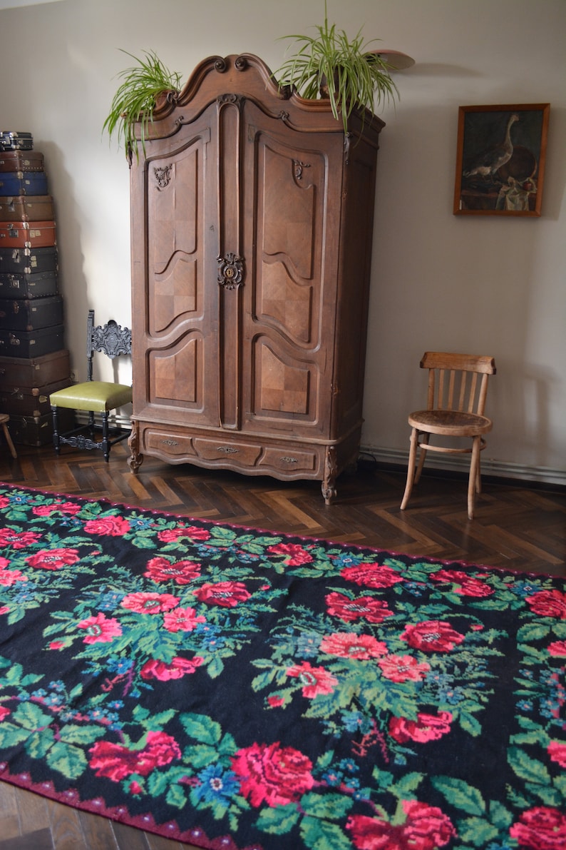 May include: A black and red floral patterned rug with green leaves. The rug is in front of a wooden armoire with two doors and a wooden chair.
