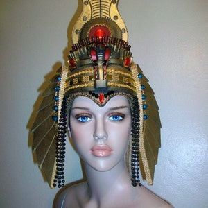 May include: A gold and black headpiece with a large, ornate crown and dangling beads. The crown features a red jewel and gold accents. The headpiece is worn by a mannequin with blue eyes.