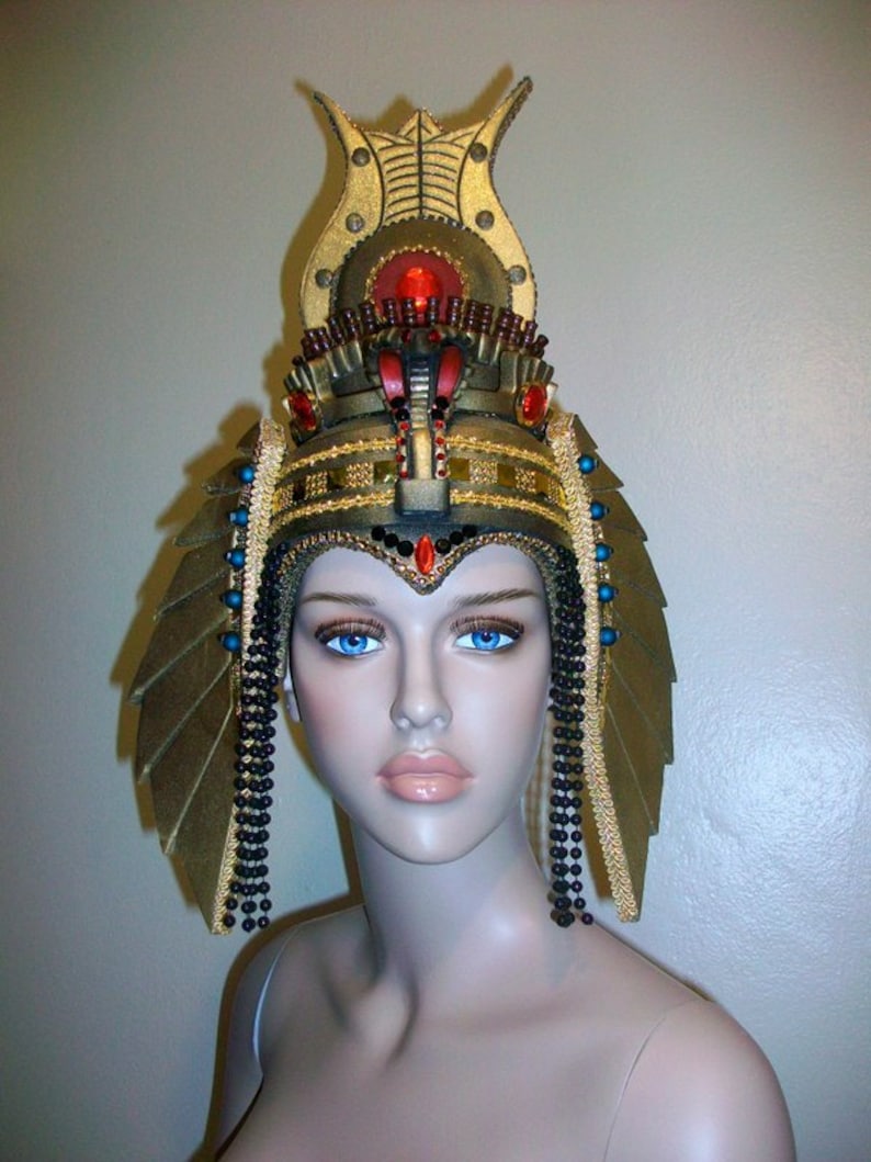 May include: A gold and black headpiece with a large, ornate crown and dangling beads. The crown features a red jewel and gold accents. The headpiece is worn by a mannequin with blue eyes.