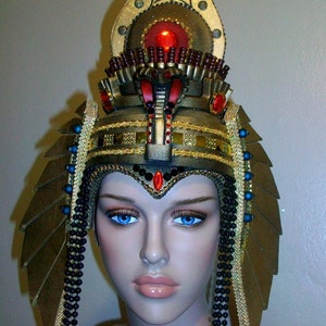 May include: A gold and black costume headpiece with a red jewel and black beads. The headpiece is designed to resemble an Egyptian pharaoh's crown.