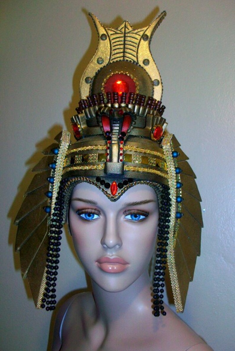 May include: A gold and black costume headpiece with a red jewel and black beads. The headpiece is designed to resemble an Egyptian pharaoh's crown.