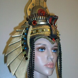 May include: A gold and black headdress with a large, ornate crown and long beaded strands. The crown features a red jewel and intricate details.