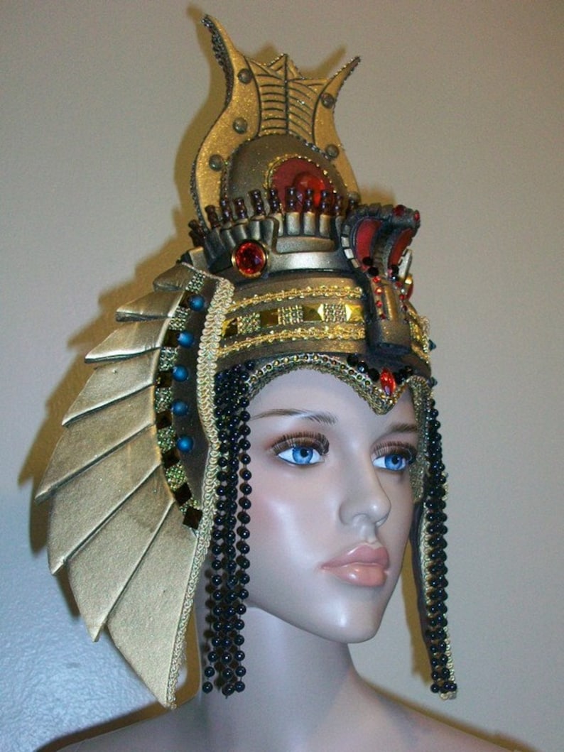 May include: A gold and black headdress with a large, ornate crown and long beaded strands. The crown features a red jewel and intricate details.