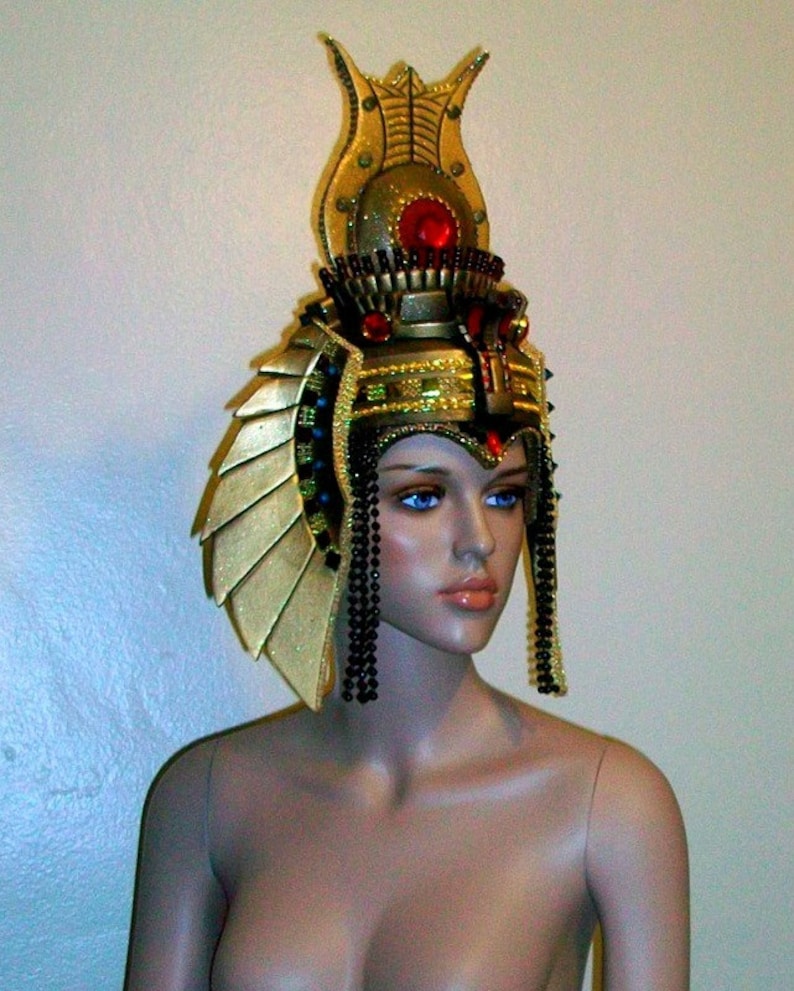 May include: A gold and black headdress with a red jewel on top. The headdress is worn by a mannequin with a neutral expression.