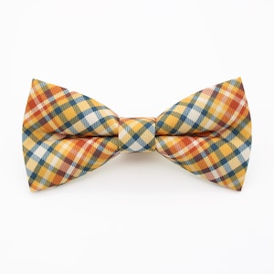 May include: A bow tie with a plaid pattern in yellow, orange, blue, and white. The bow tie is set against a white backdrop. The fabric features a traditional plaid design, suitable for various occasions.