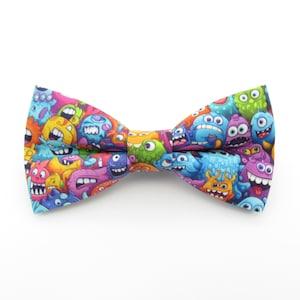 Può includere: Un papillon colorato con un motivo di mostri dei cartoni animati in vari colori, tra cui blu, verde, viola, arancione e giallo.