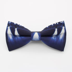 May include: A navy blue bow tie featuring a stage lighting design. The bow tie has a repeating pattern of white spotlights against a dark blue background. The bow tie is a classic accessory for formal events.