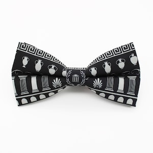 May include: A black and white bow tie featuring a Greek-inspired design. The bow tie has a repeating pattern of white vases, columns, and geometric shapes against a black background. The center of the bow tie features a building with a laurel wreath.