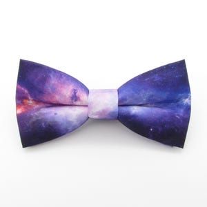 Puede incluir: Una pajarita de color morado y rosa con estampado de galaxia. La pajarita está hecha de una tela suave y cómoda y tiene una forma clásica de pajarita.