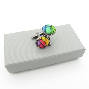 Pode incluir: Um par de botões de punho numa caixa cinzenta. Os botões de punho são de prata com desenhos redondos, coloridos, como vitrais.