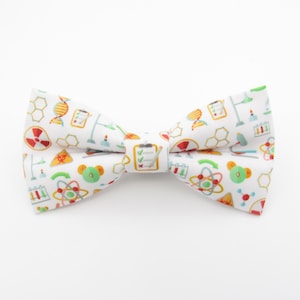 May include: White bow tie with a science-themed pattern. The pattern includes images of beakers, test tubes, atoms, and other science-related symbols.