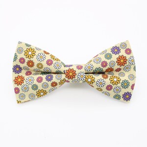 May include: A light beige bow tie with a repeating floral pattern in orange, yellow, purple, white, and green. The bow tie is a classic shape, suitable for formal or casual wear.